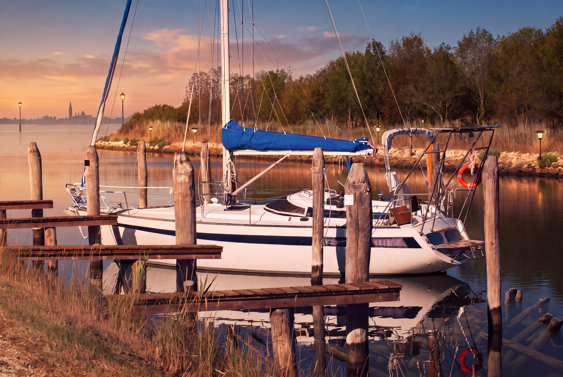 Sailboat moored at wooden poles in a calm lagoon at sunset with a distant bell tower