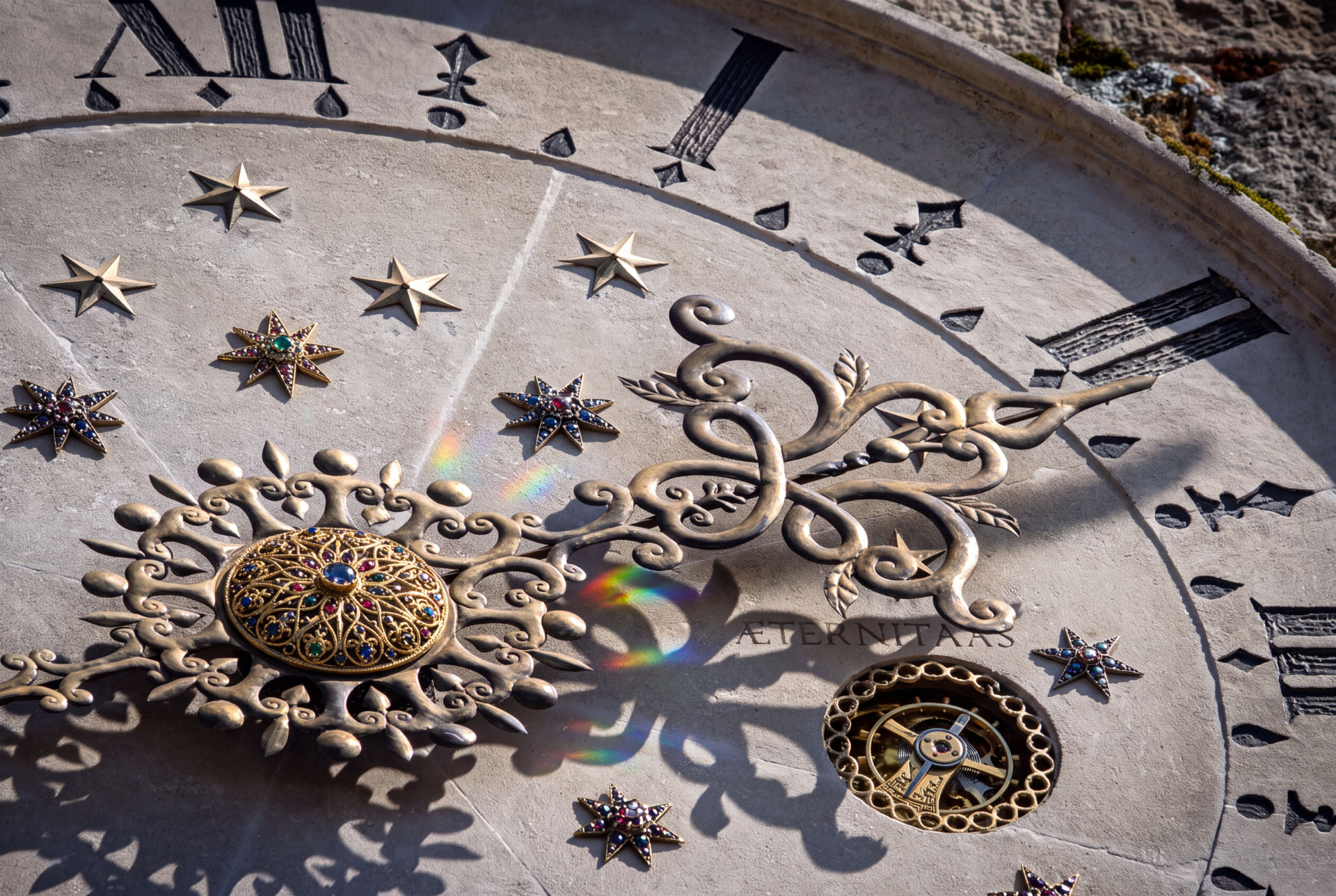 Close-up of an ornate astronomical clock face with gold hands, jewelled stars and Roman numerals casting shadows and small rainbows on the stone
