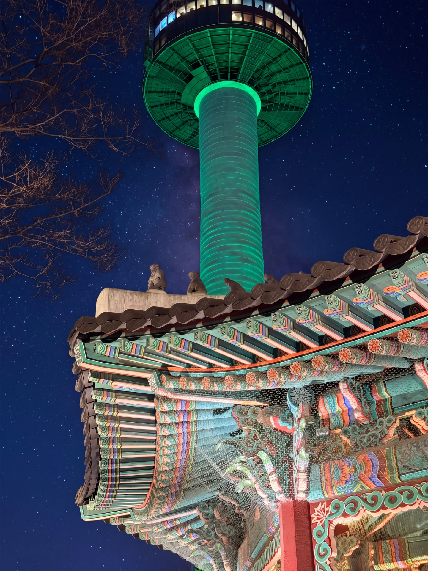 Green-lit Seoul tower rising behind a traditional curved tiled roof at night