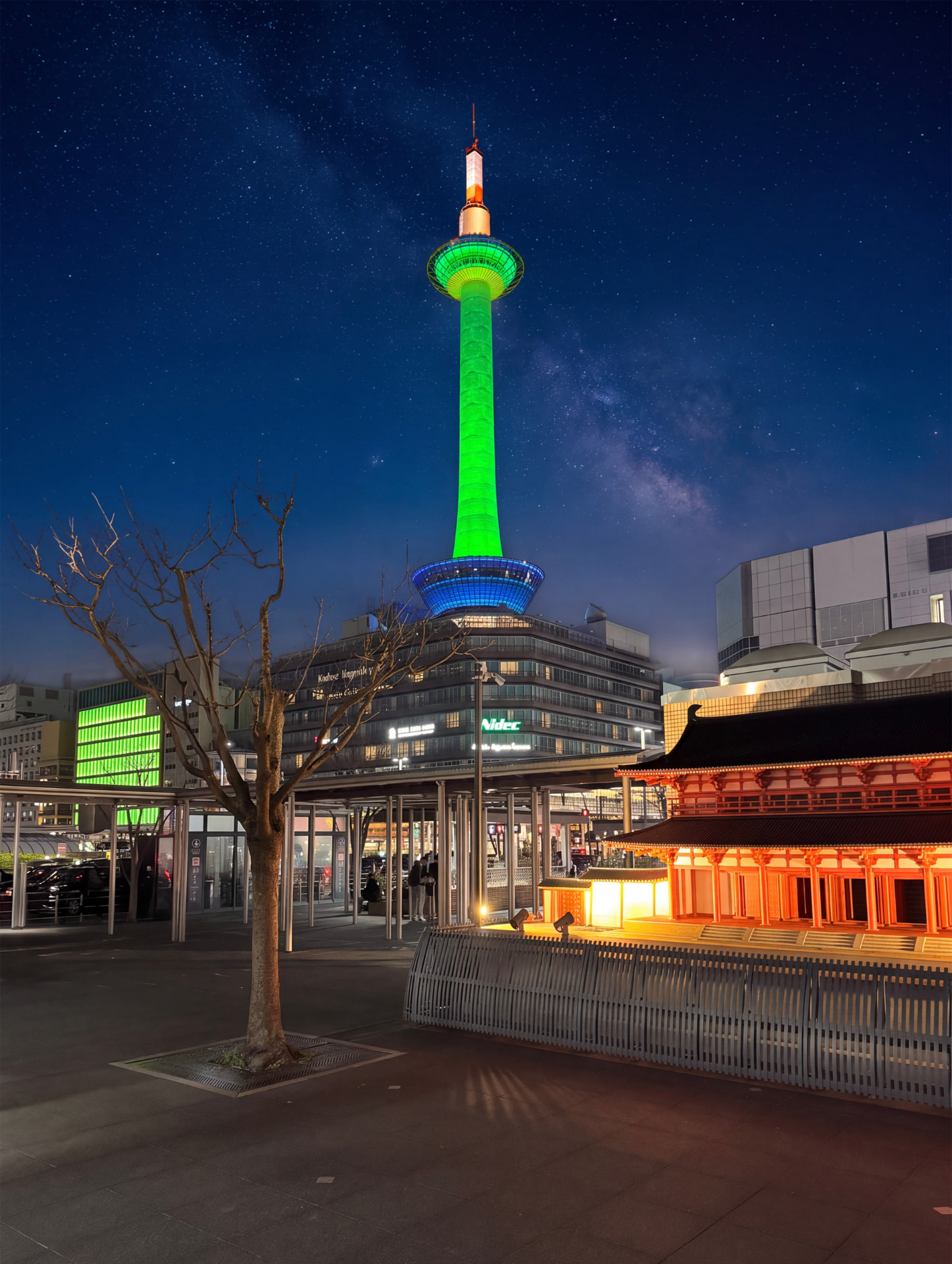 Kyoto Tower lit in green at night above a red-lit temple building