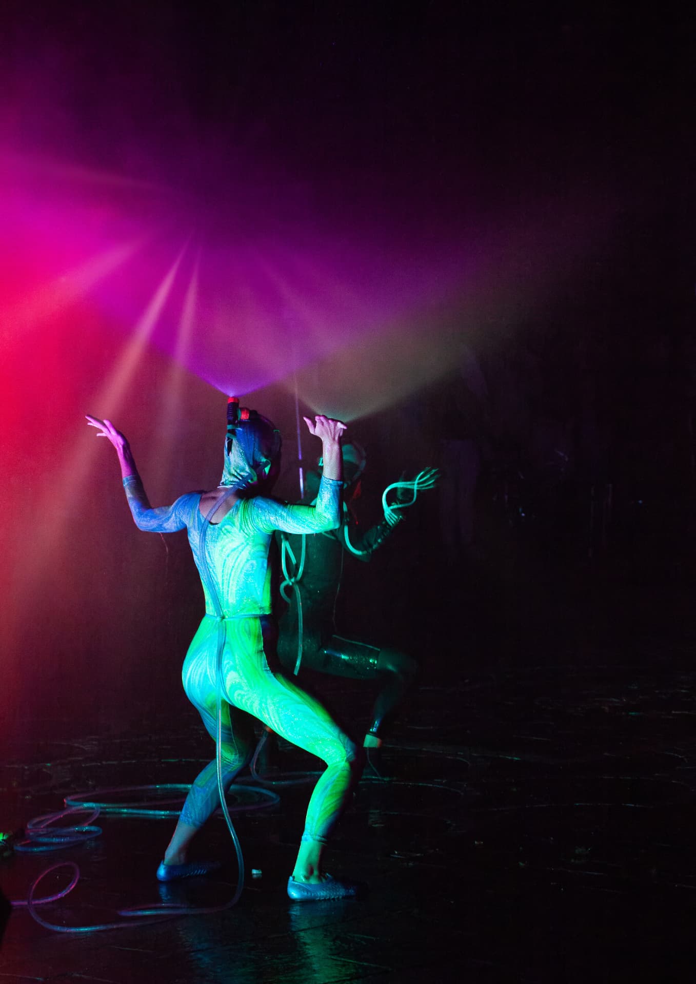 Two performers in glowing green costumes dancing in coloured mist and pink and purple light