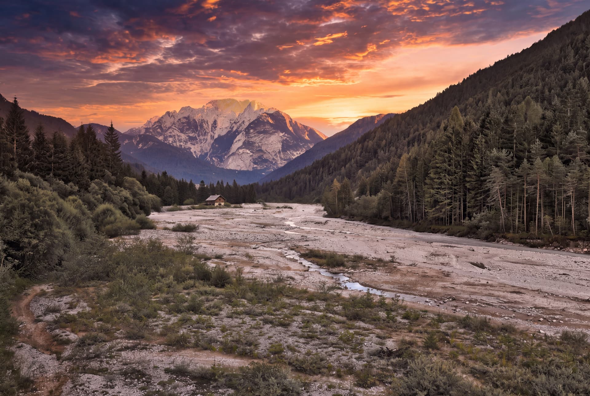 Alpine valley with a gravel riverbed, a small wooden hut and a rocky mountain lit by an orange sunset sky