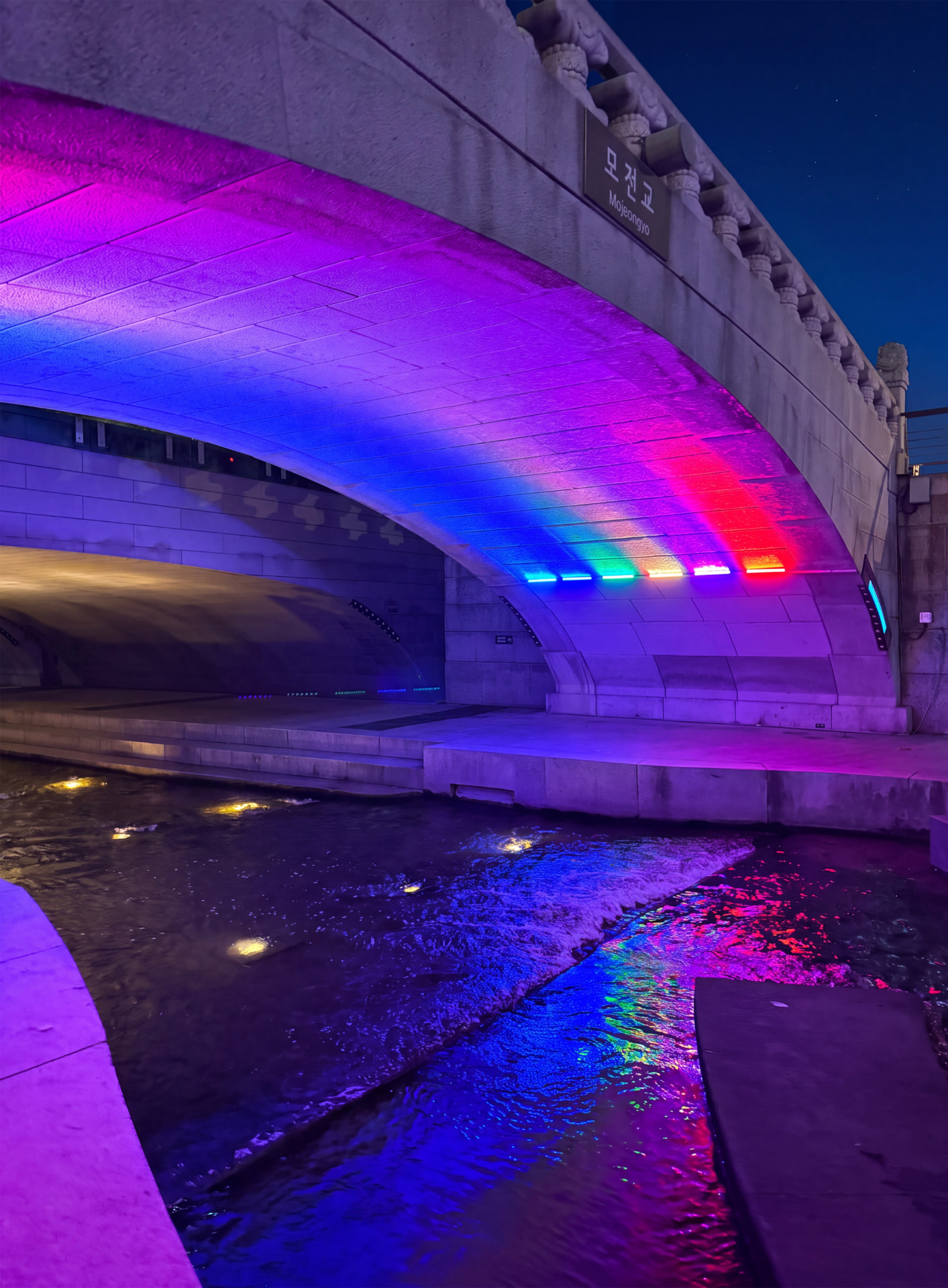 Bridge arch lit in purple and blue neon above a stream at night