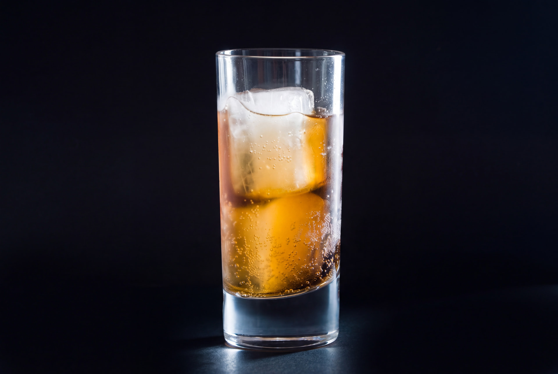 Tall glass of sparkling amber drink with ice cubes against a black background