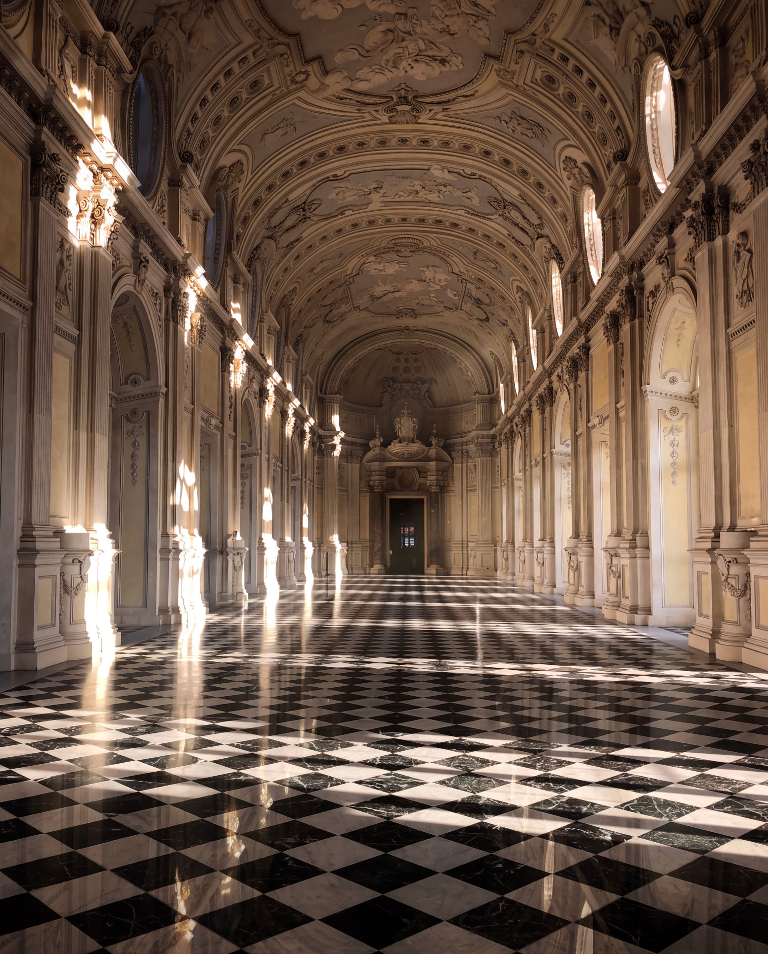 Long gallery of the Reggia di Venaria Reale with a black and white checkered floor, arches and light reflecting from the windows