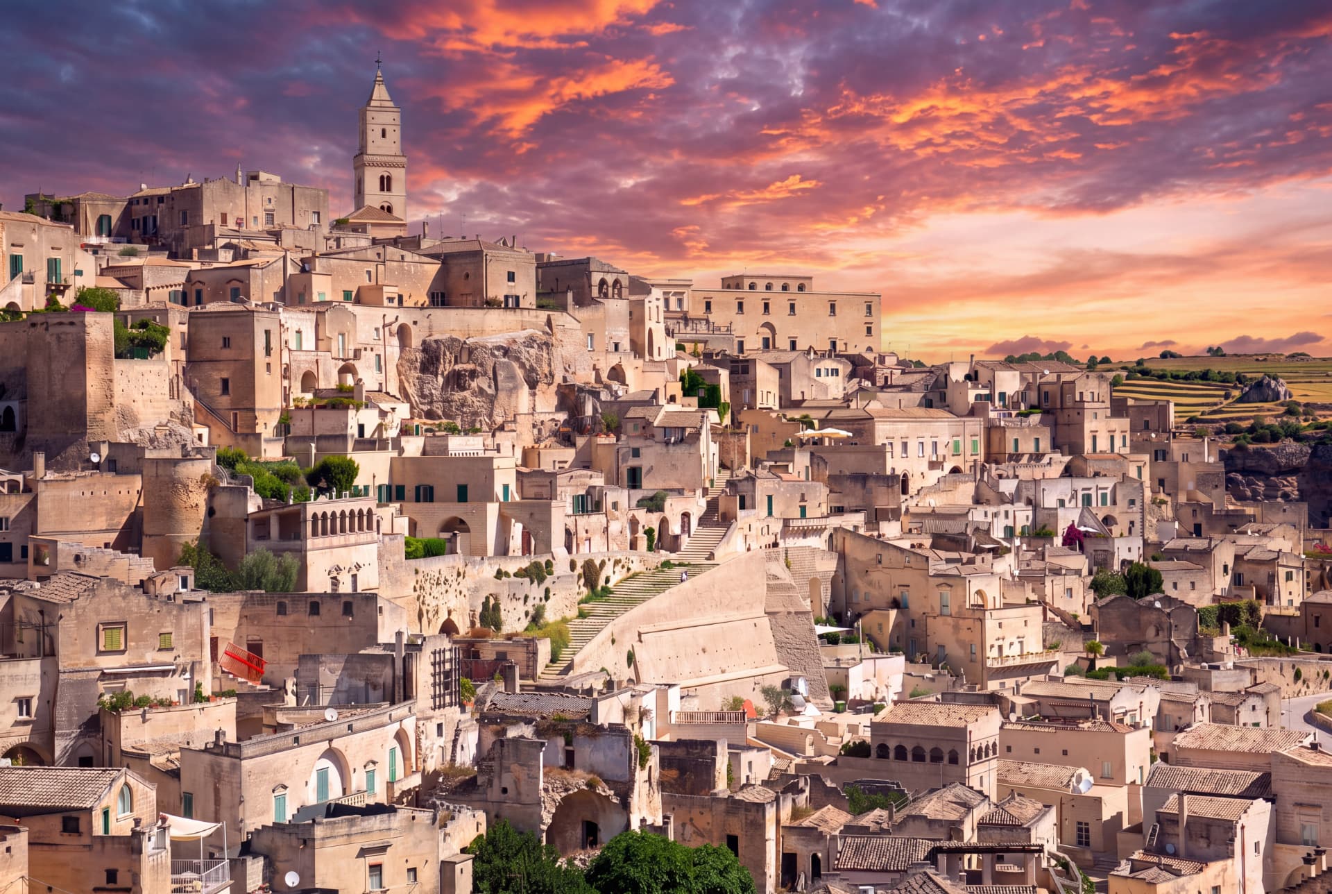 Hillside old town of Matera with stone houses and a bell tower under an orange and purple sunset sky
