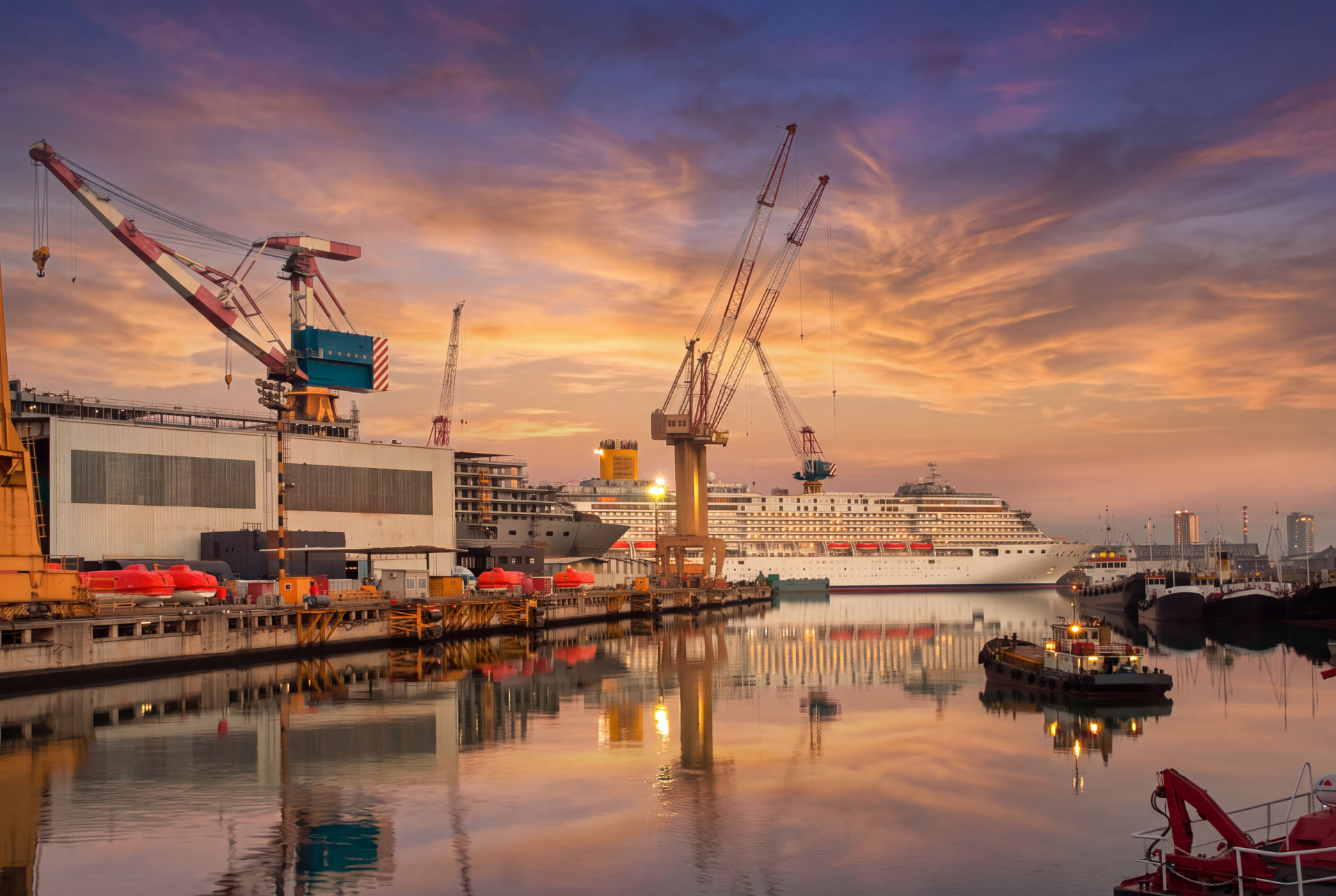 Shipyard cranes and a cruise ship reflected in calm water under an orange and purple sunset sky